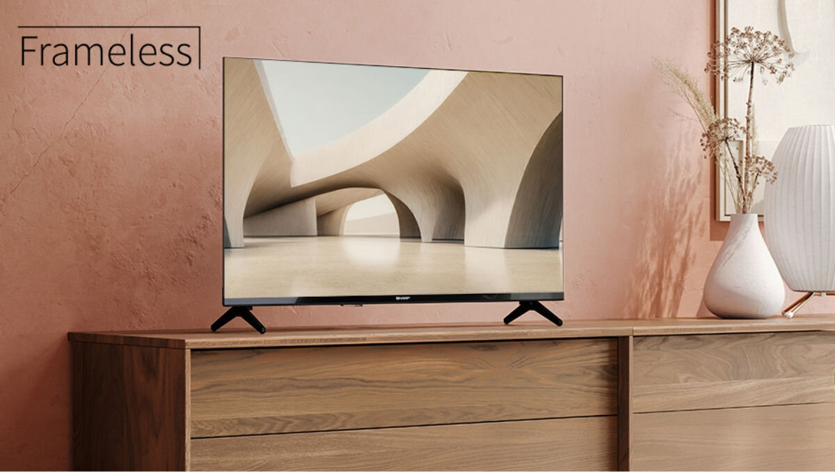 Telewizor z cienkimi ramkami stoi na drewnianej komodzie, a na ekranie widoczna jest nowoczesna architektura. Obok telewizora znajduje się wazon z suchymi roślinami, a na ścianie widnieje napis \'Frameless\'.